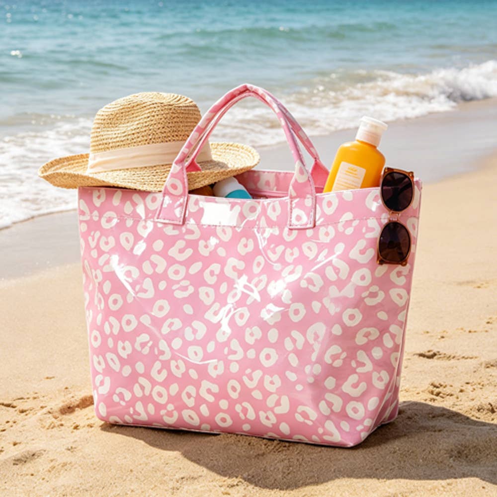 A pink leopard-print tote bag on a sandy beach with a straw hat, sunglasses, and sunscreen in it, with the ocean in the background.