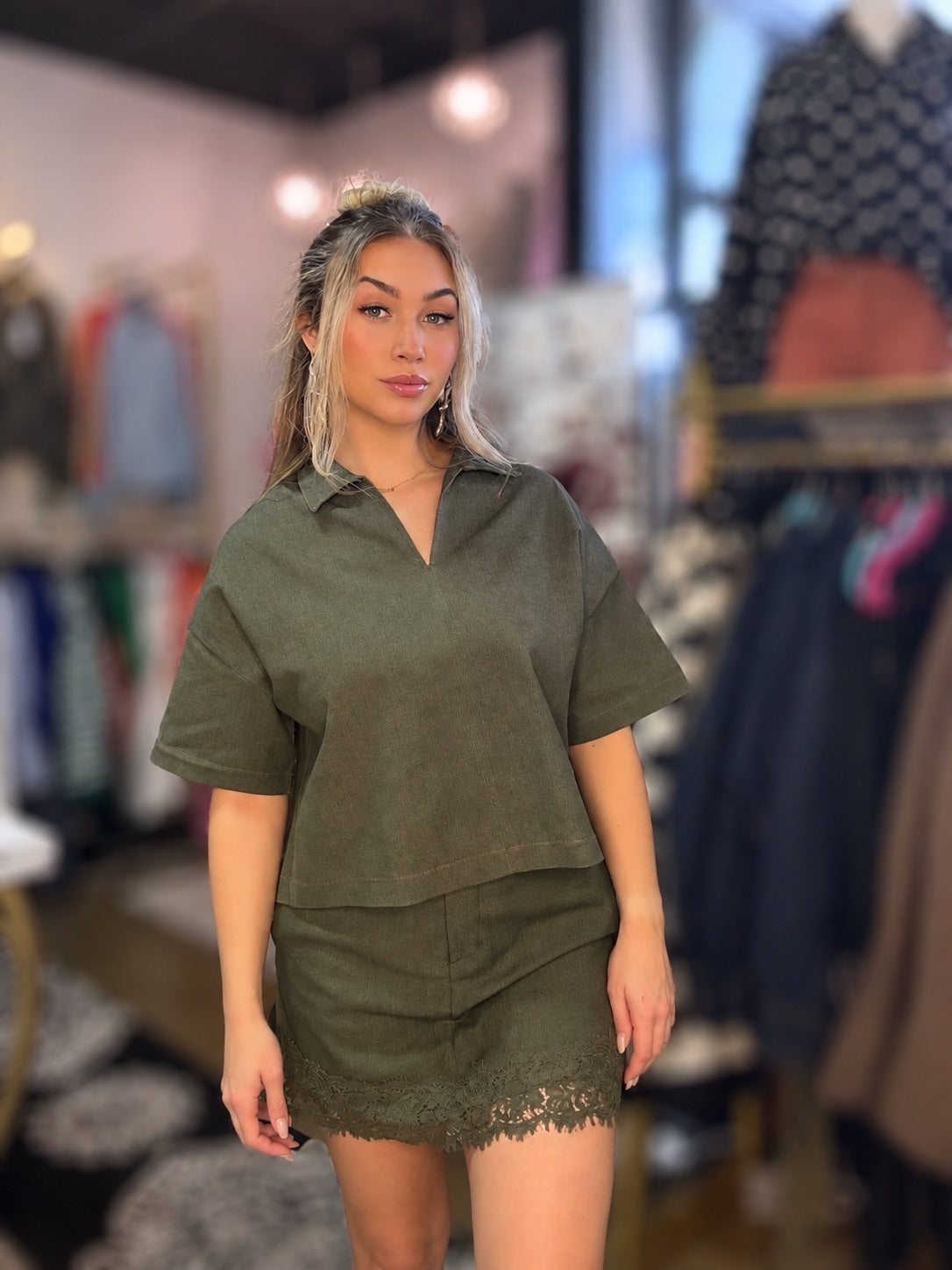 A woman modeling an olive green two-piece set consisting of a short-sleeve collared top and a matching mini skirt with a lace hem, set against a blurred boutique background.