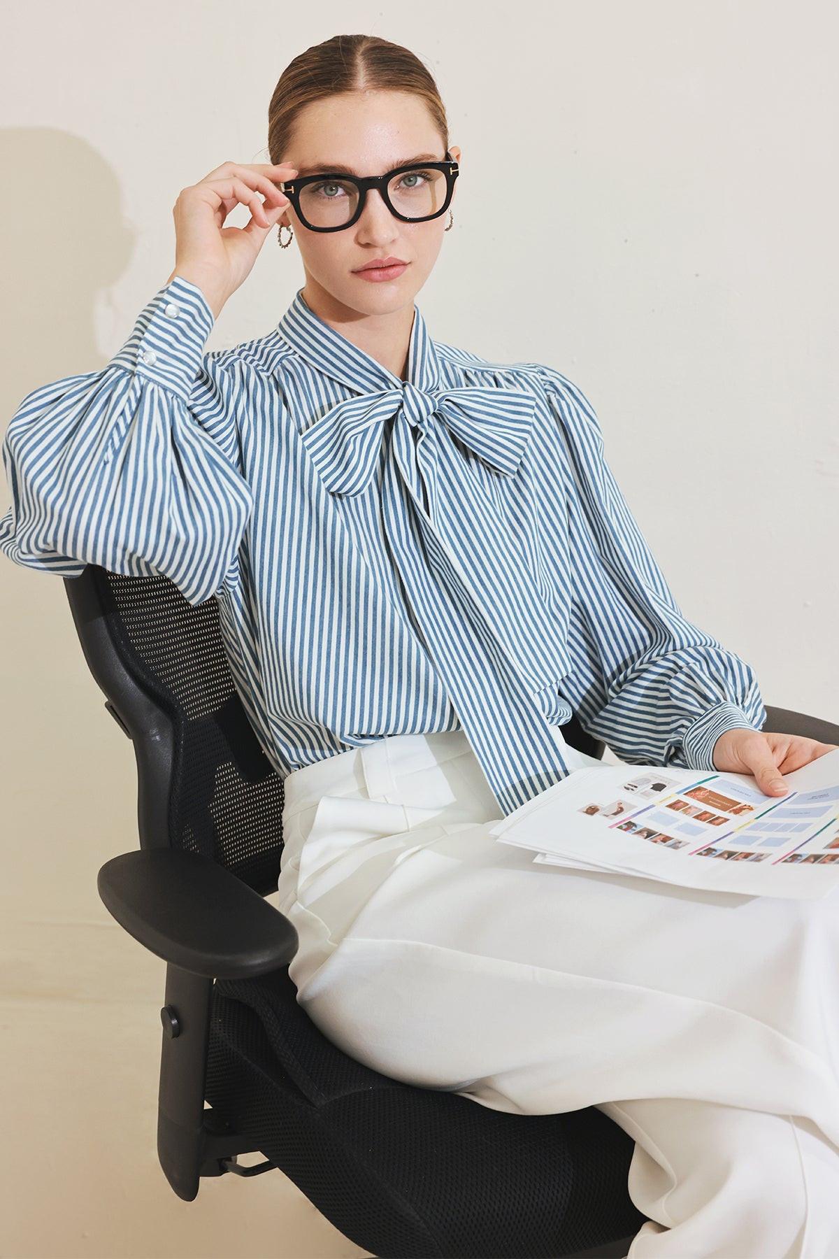 Person sitting in a chair wearing a blue striped shirt with a bow tie, holding a magazine.