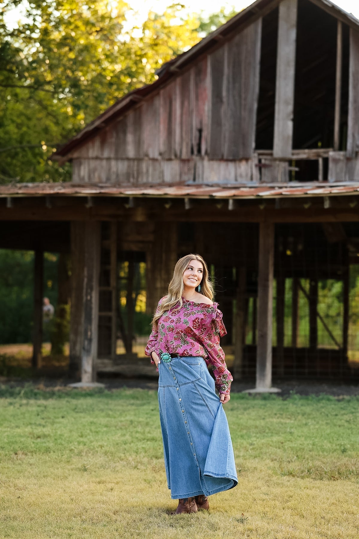 A woman stands in a grassy field in front of a rustic wooden barn, wearing a floral off-the-shoulder long-sleeve top and a long blue denim skirt.