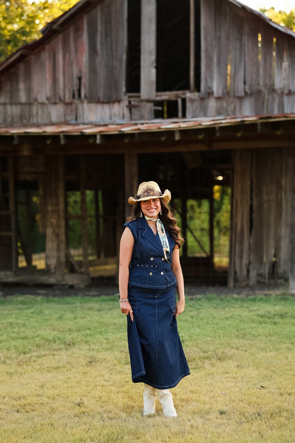 A woman stands in a grassy field in front of a rustic wooden barn, wearing a navy blue denim vest and matching long skirt set, a tan cowboy hat, and white boots.