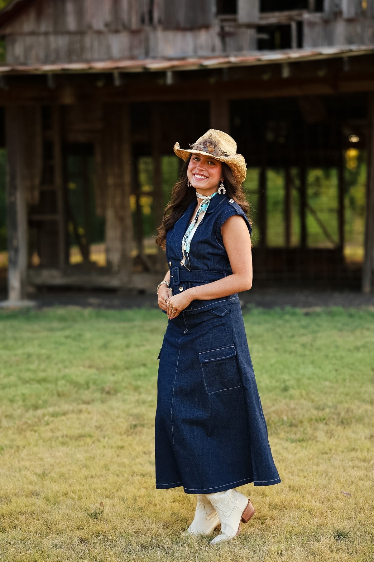 A woman stands outdoors in front of a rustic wooden barn on a grassy field, wearing a sleeveless dark blue denim dress with a cowboy hat, white boots, and a white neck scarf.