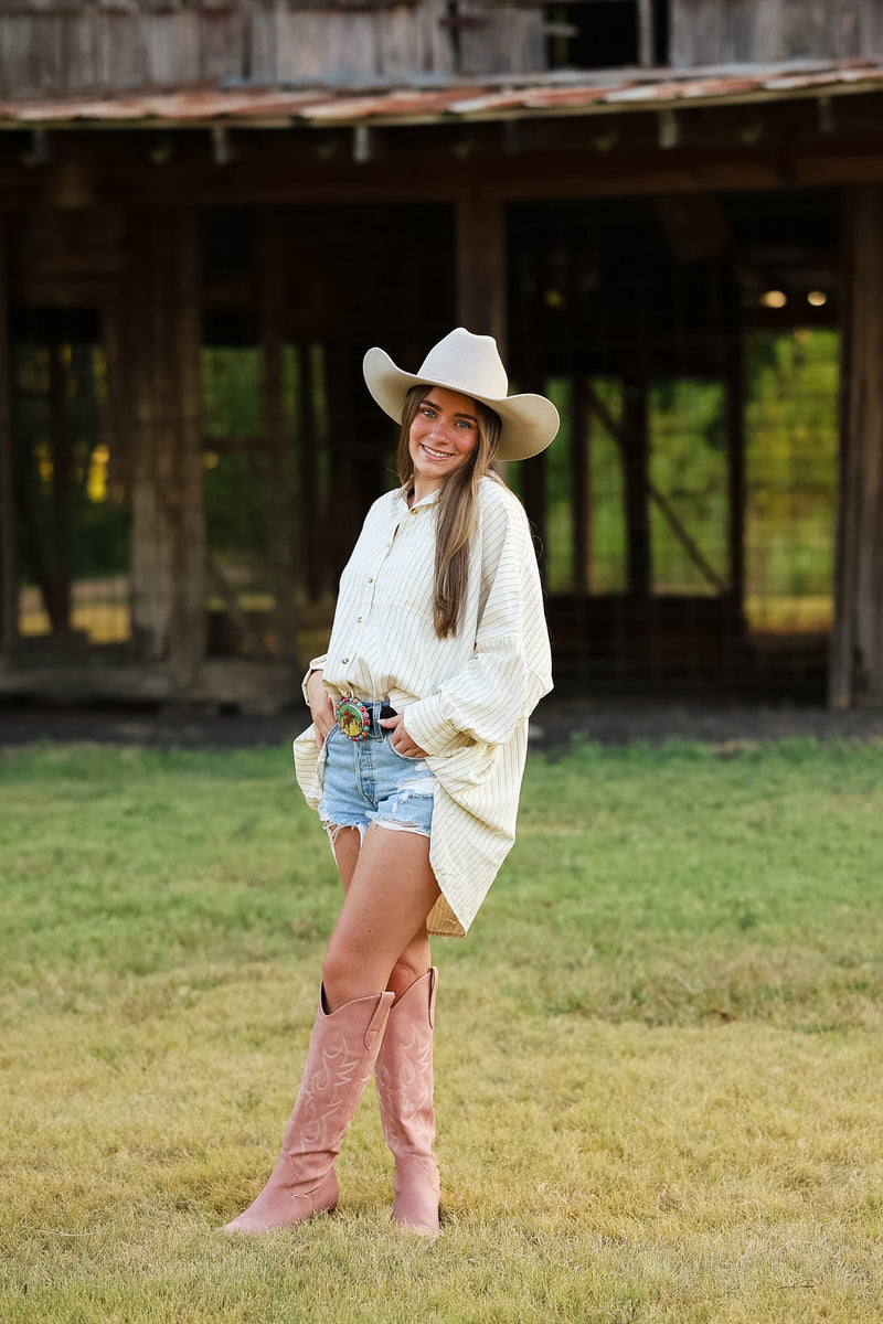 A woman stands outdoors in front of a rustic wooden barn wearing a cream ribbed knit poncho, denim shorts, a cream cowboy hat, and pink cowboy boots.
