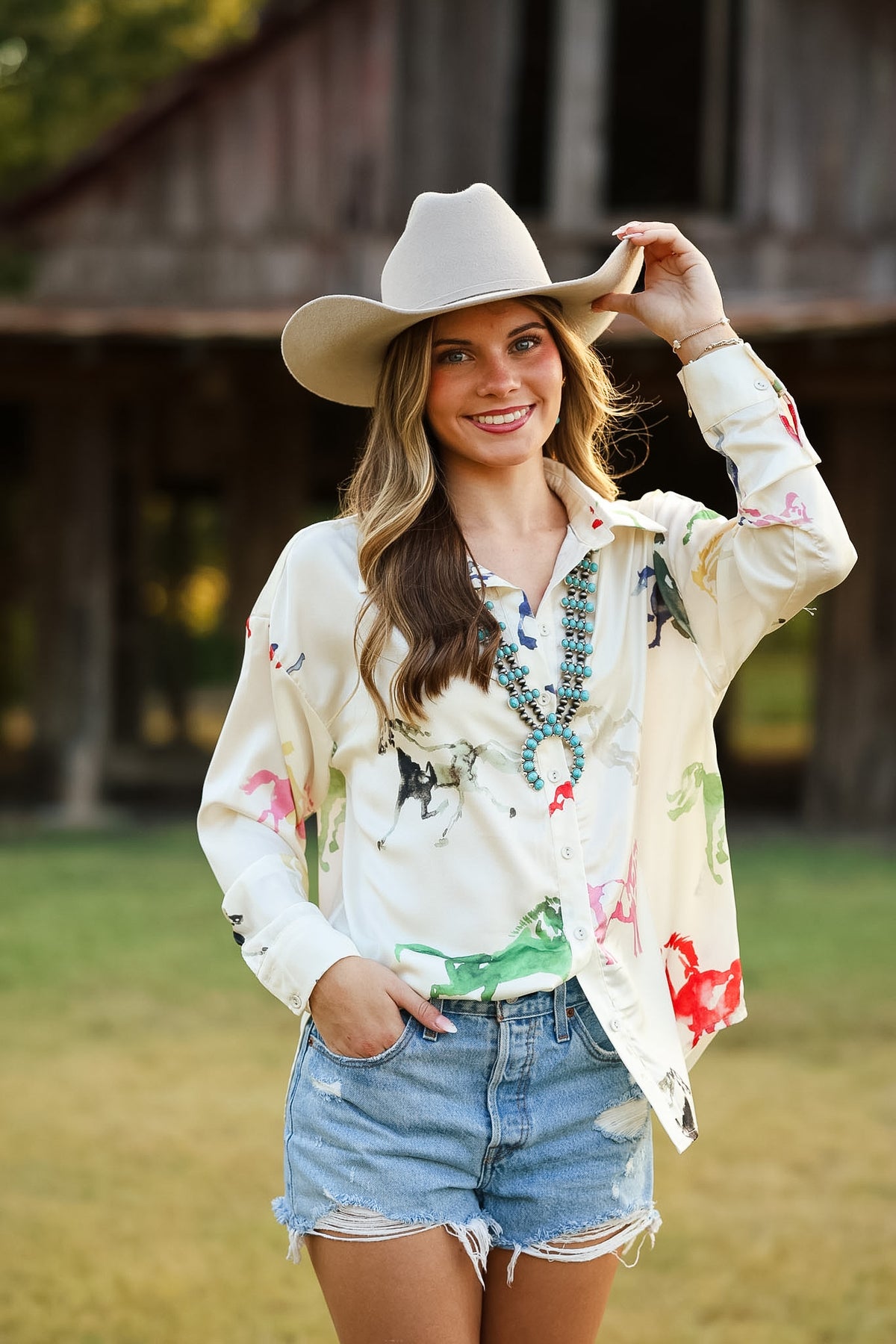 A woman wearing a cream cowboy hat, a cream western-style button-up shirt with colorful horse prints, and light-wash denim shorts, standing outdoors in front of a rustic wooden barn.