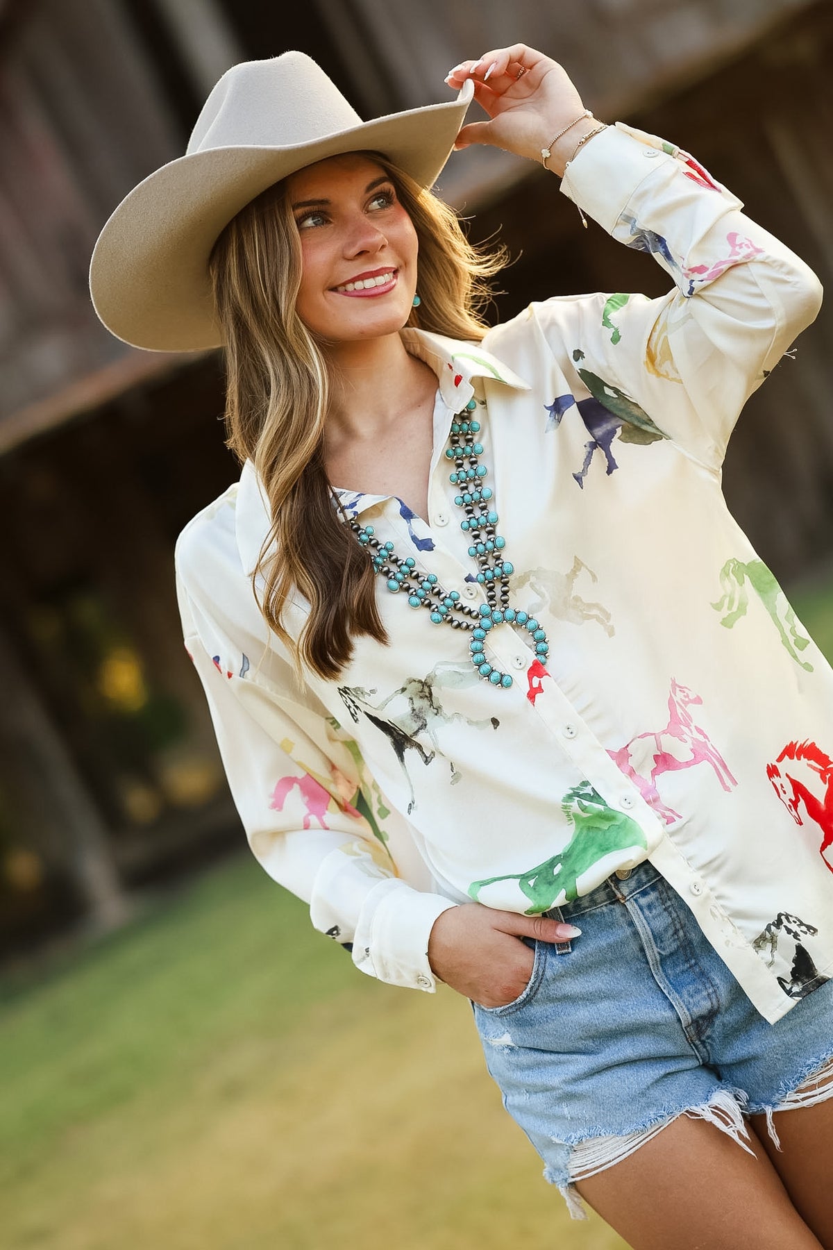 A woman wearing a cream western-style button-up shirt with colorful horse prints, blue denim shorts, a turquoise beaded necklace, and a beige cowboy hat, posing outdoors in front of a rustic wooden barn.