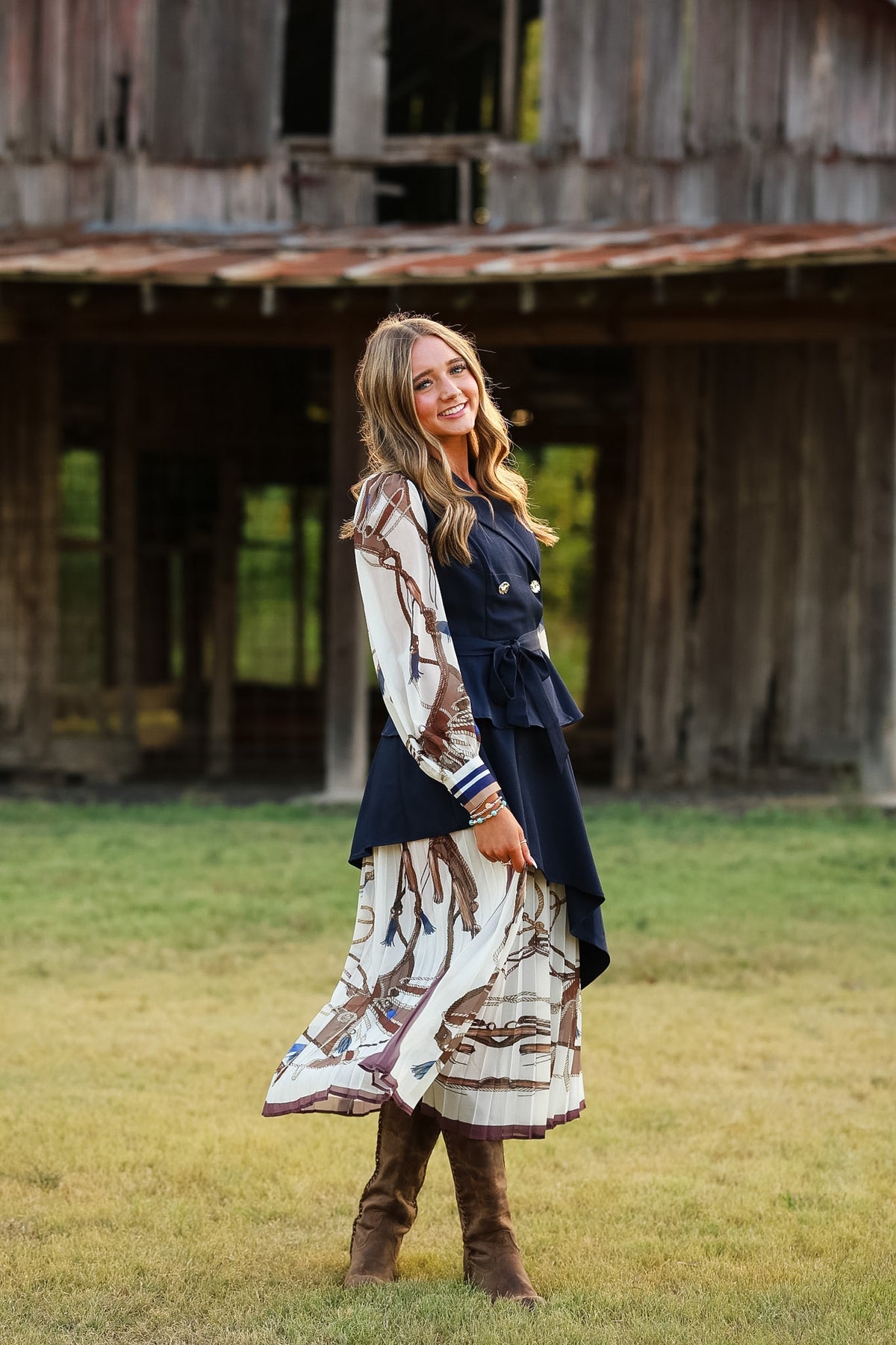 A woman stands outdoors in front of a rustic wooden barn, wearing a long-sleeve navy vest over a cream and brown patterned dress with brown knee-high boots.