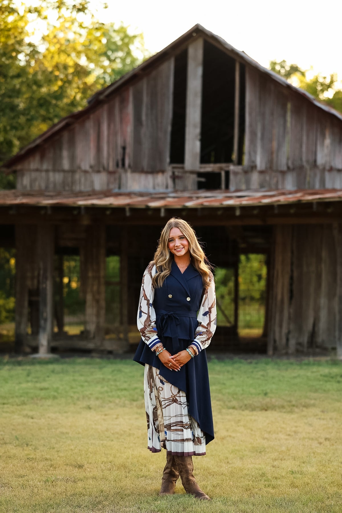 A woman stands in a grassy field in front of a rustic wooden barn, wearing a navy blue double-breasted vest over a long-sleeve patterned dress with brown boots.