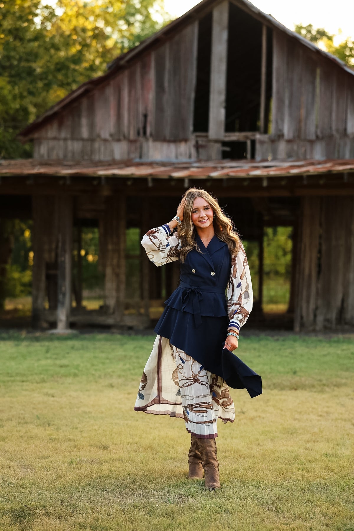 A woman stands in a grassy field in front of a rustic wooden barn, wearing a navy blue double-breasted vest over a long-sleeve cream and brown patterned dress, paired with brown boots.