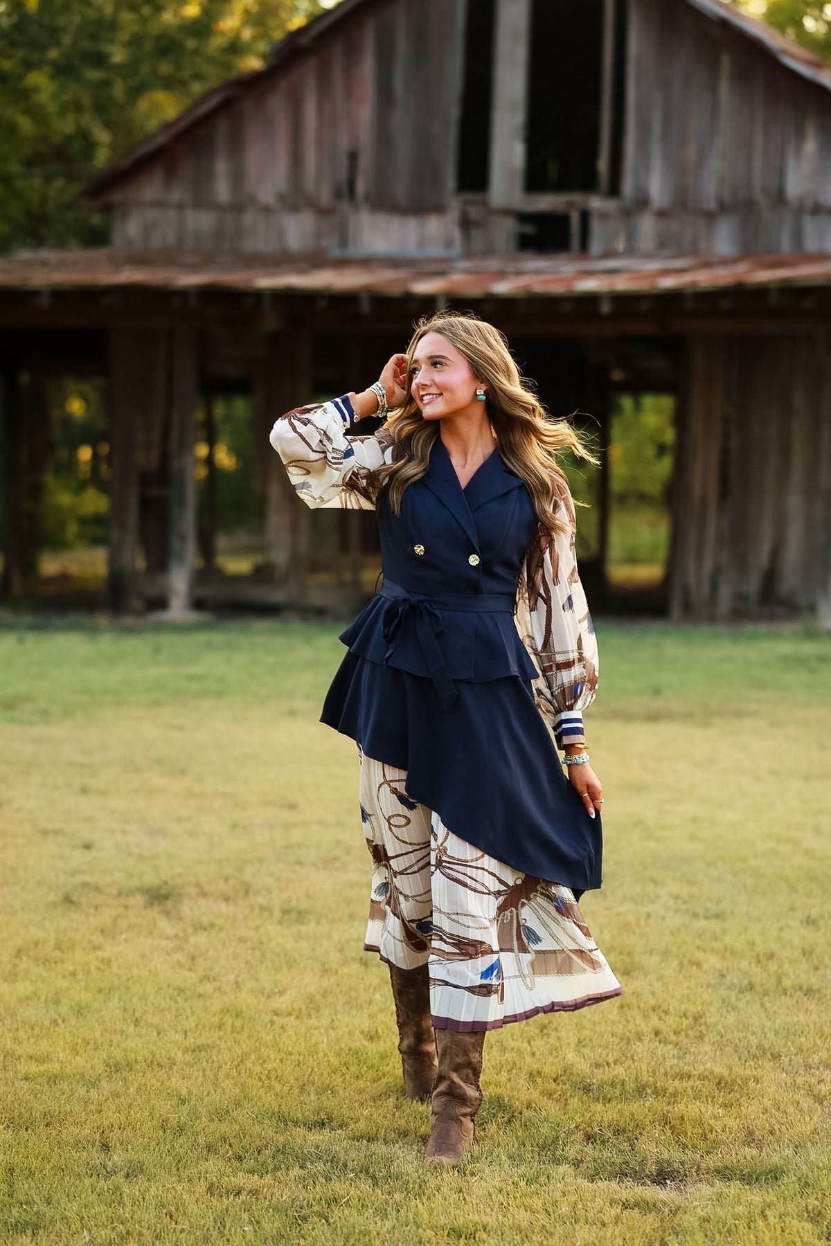A woman stands in a grassy field in front of a rustic wooden barn, wearing a navy blue belted vest over a cream and brown patterned long-sleeve top and matching wide-leg pants, paired with brown boots.