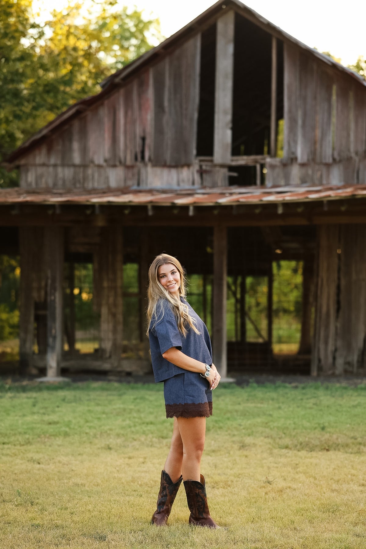 A woman stands in a grassy field in front of a large rustic wooden barn, wearing a blue short-sleeve top and skirt set with dark brown cowboy boots.