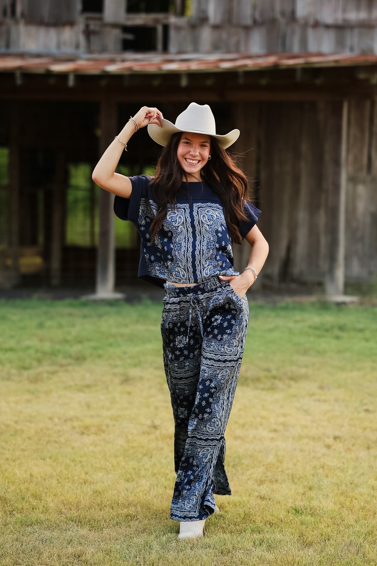 A woman stands outdoors in a grassy field wearing a matching navy and white paisley bandana set, consisting of a short-sleeve crop top and wide-leg pants, along with a white cowboy hat.