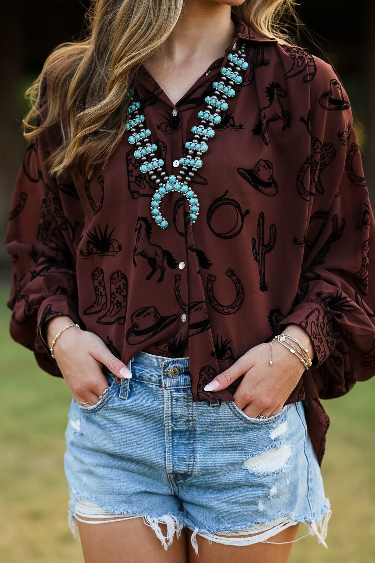 A woman wearing a brown long-sleeve button-up shirt with a black western-themed print of hats, boots, and cacti, paired with light-wash denim shorts and a large turquoise statement necklace.