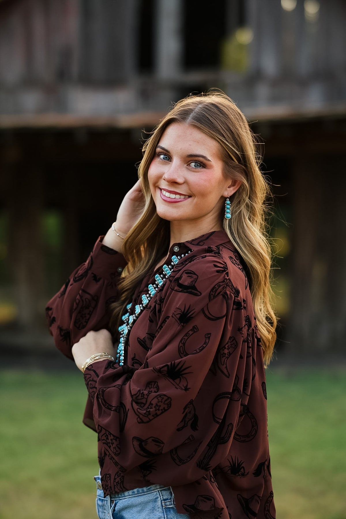 A woman wearing a dark brown long-sleeve blouse with a black horseshoe and cattle print, styled with blue jeans and turquoise jewelry, standing outdoors in front of a rustic wooden structure.