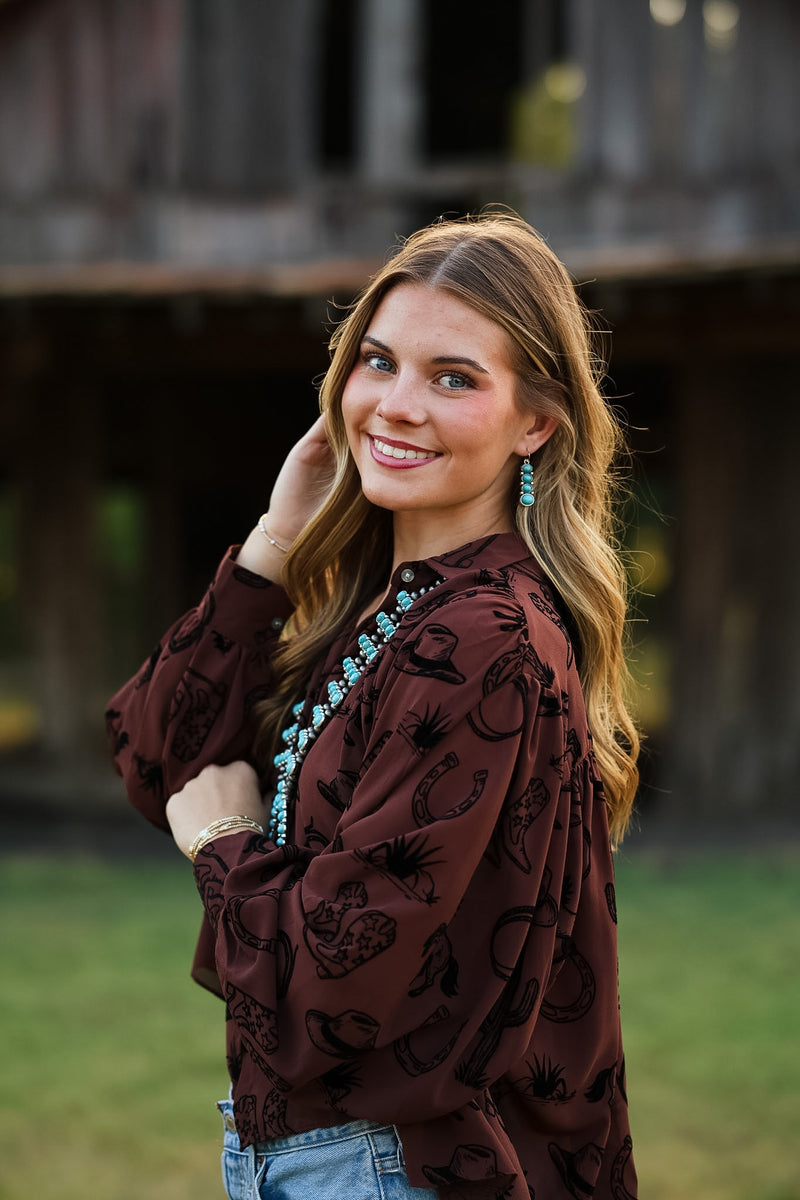 A woman wearing a dark brown long-sleeve blouse with a black horseshoe and cattle print, styled with blue jeans and turquoise jewelry, standing outdoors in front of a rustic wooden structure.