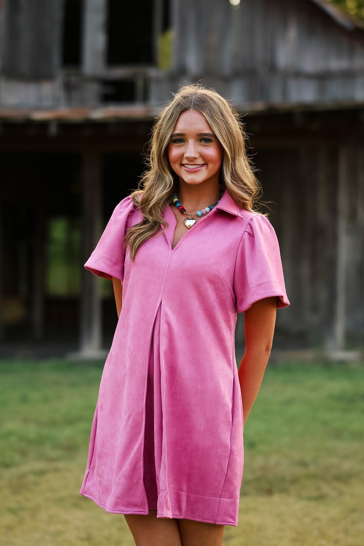A woman wearing a short-sleeve pink collared dress stands outdoors in front of a rustic wooden barn and grassy field.