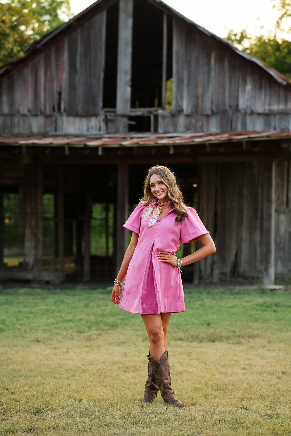 A woman stands in a grassy field in front of a rustic wooden barn, wearing a short-sleeve pink mini dress and brown cowboy boots.