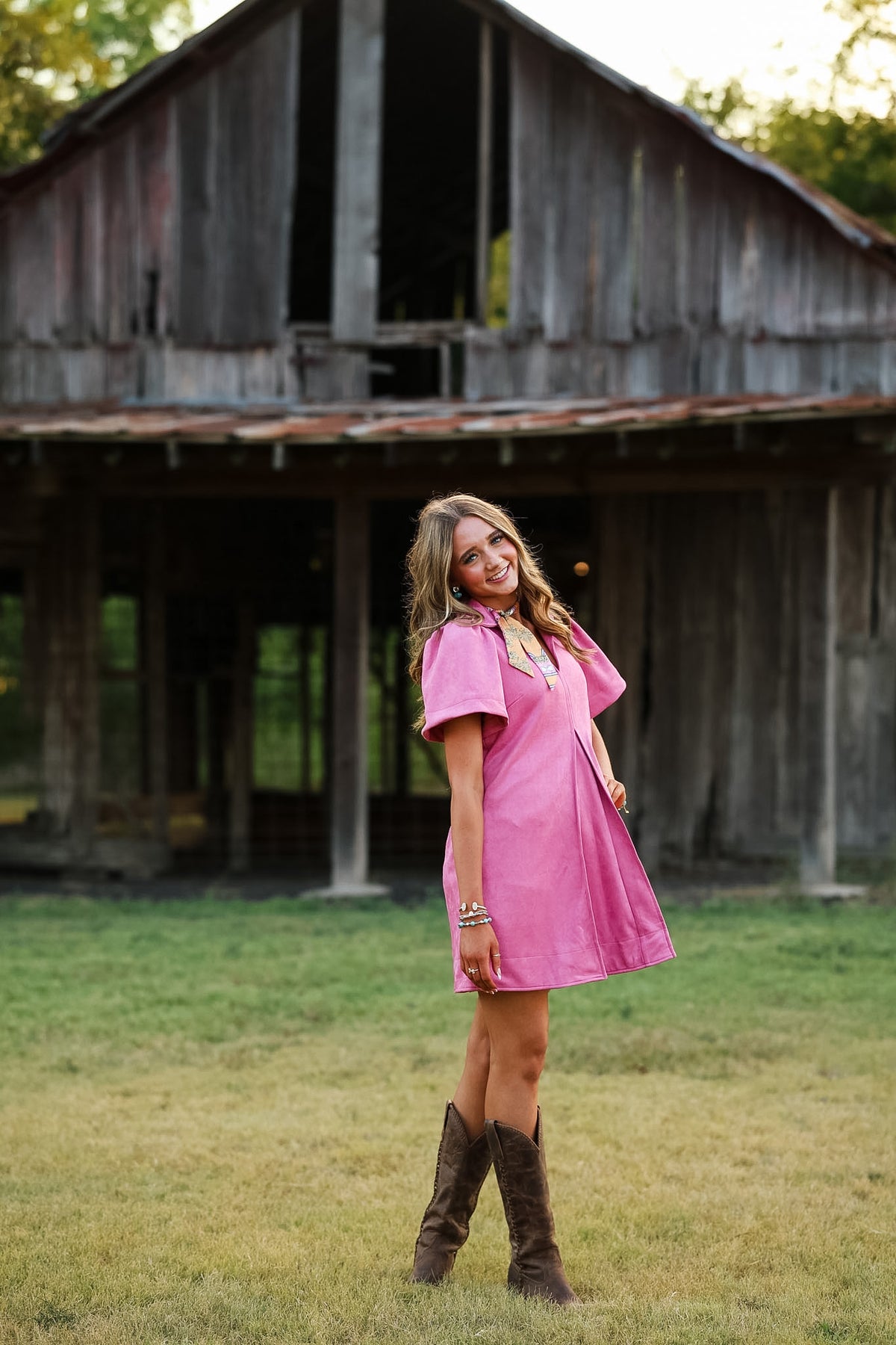 A woman wearing a pink short-sleeve mini dress and brown cowboy boots stands in a grassy field with a weathered wooden barn in the background.