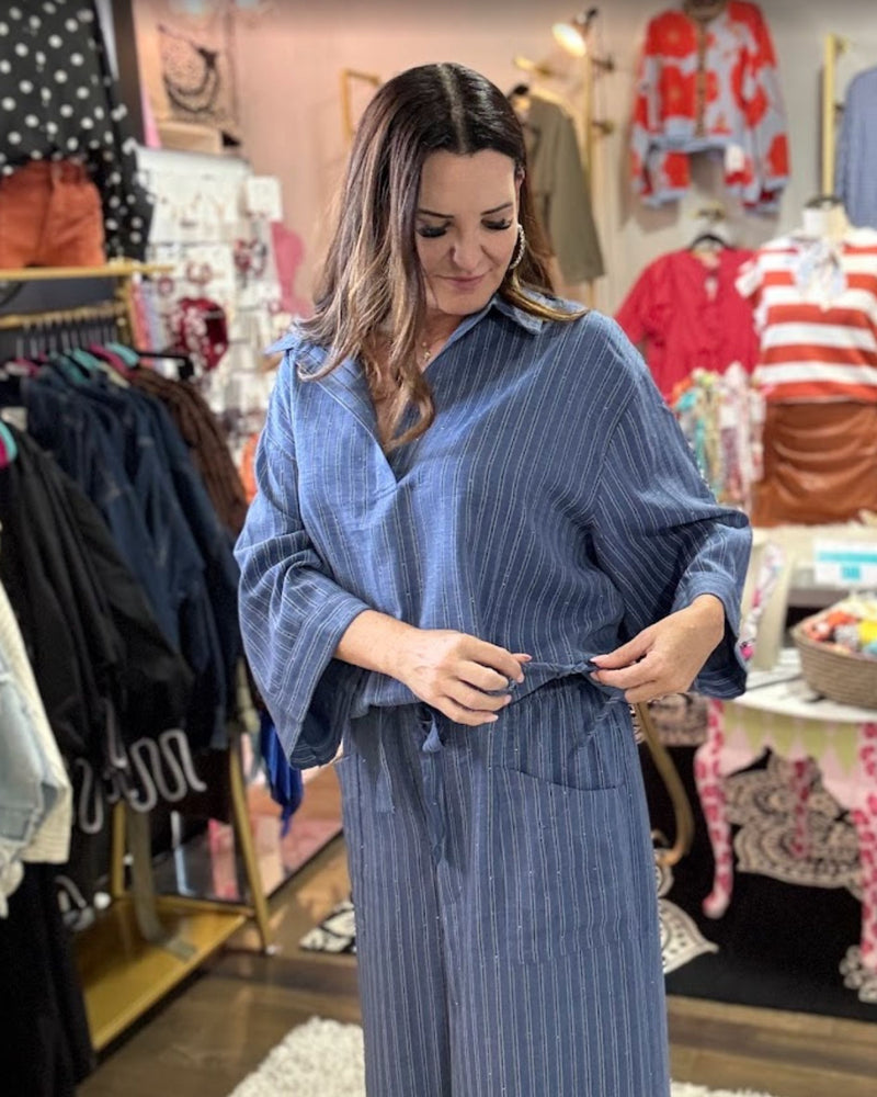 A woman modeling a blue and white vertically striped long-sleeve shirt dress in a clothing boutique, adjusting the front waist tie.