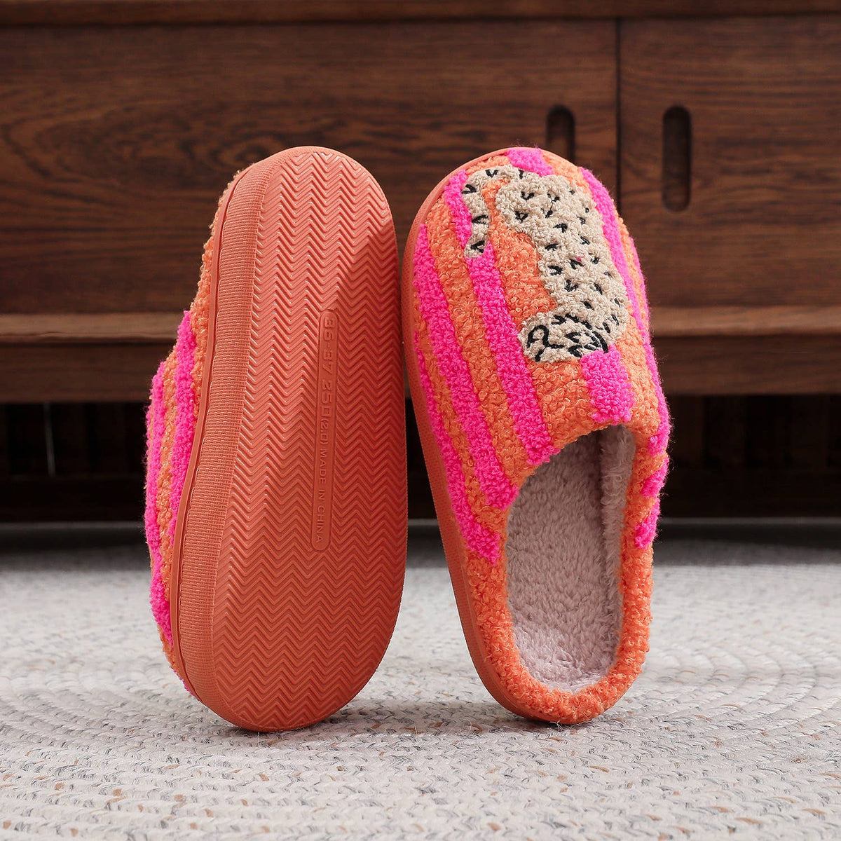 A pair of fuzzy slip-on house slippers with orange, pink, and beige striped knit-style uppers and a soft pink fleece lining, shown standing on a light rug in front of a dark wooden cabinet.