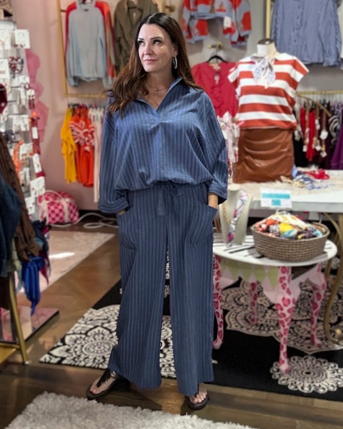 A woman stands in a clothing boutique wearing a matching blue pinstriped button-up shirt and wide-leg pants set. The store is filled with racks of colorful garments, mannequins, and display tables.