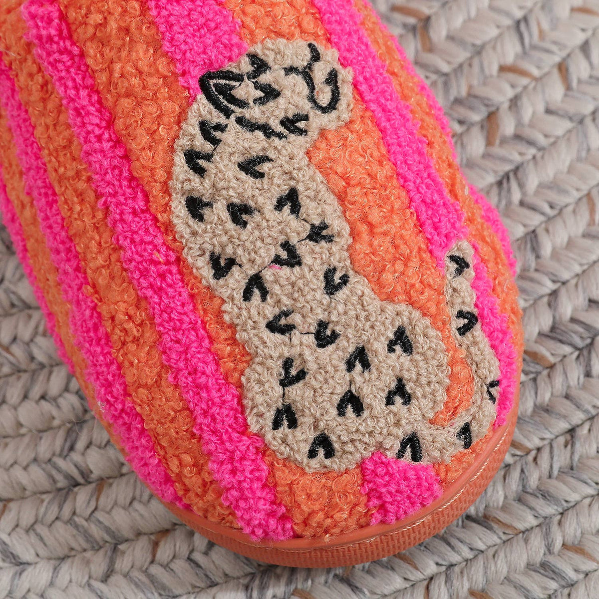 A close-up of a textured slipper featuring bright pink and orange vertical stripes, with a beige crocheted cat design outlined in black on the side, resting on a woven straw surface.