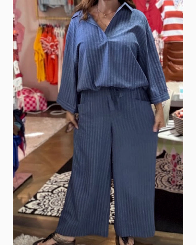 A woman wearing a blue striped two-piece set consisting of a long-sleeve collared top and matching wide-leg pants, photographed in a clothing store.