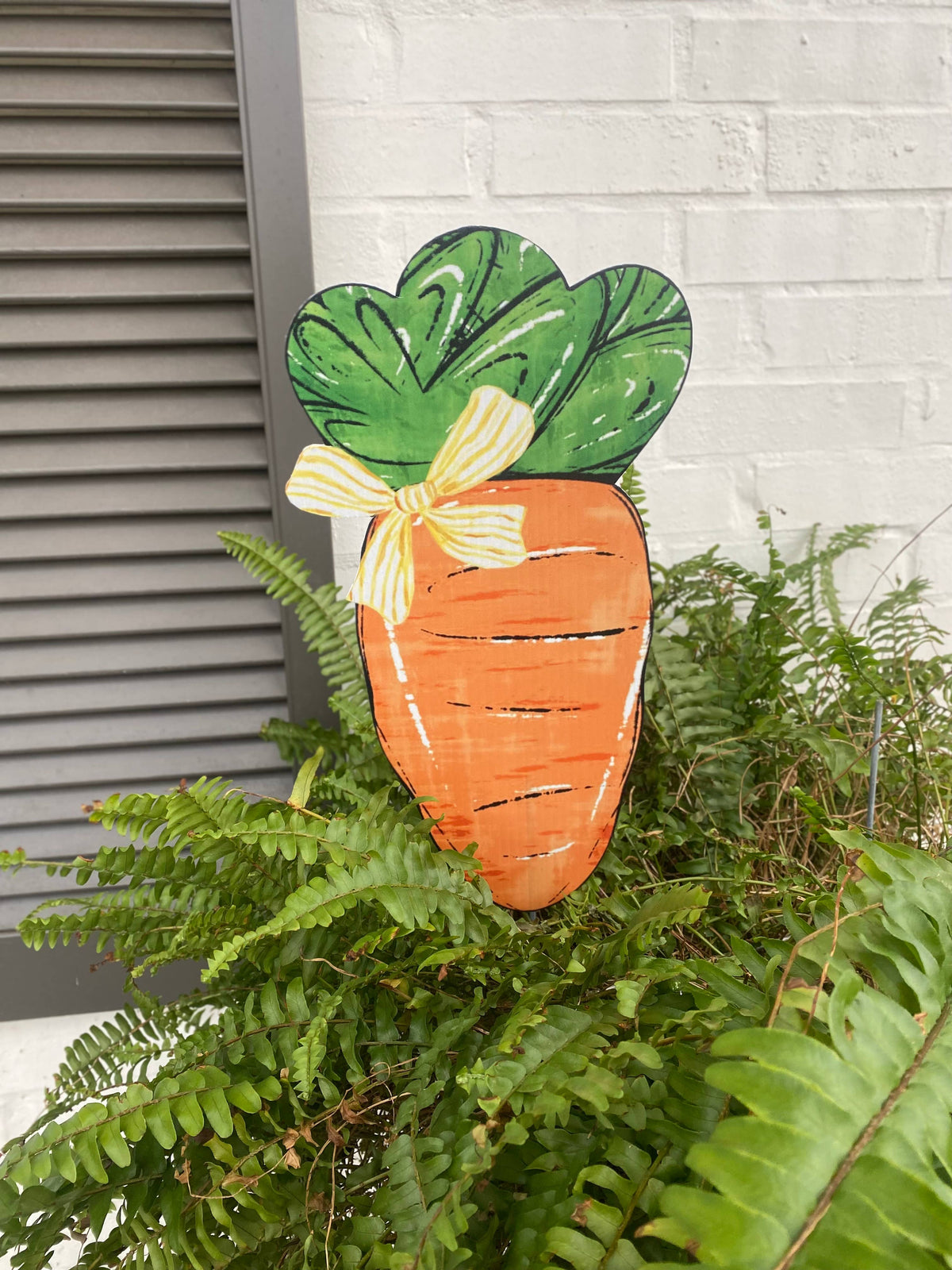Decorative carrot-shaped sign with green leaves and a yellow bow on a plant against a white wall.