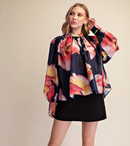 A woman modeling a long-sleeve blouse with a dark navy base and a large, vibrant floral print in pink, red, yellow, and orange, paired with a black skirt against a solid beige background.