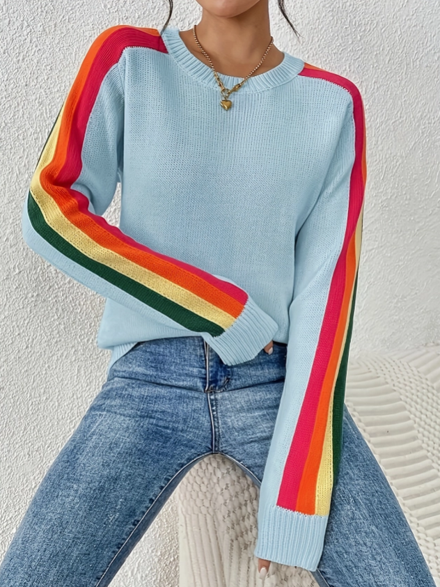 A woman wearing a light blue crewneck sweater with bold rainbow stripes running down both sleeves, paired with blue jeans, sitting against a textured white wall.