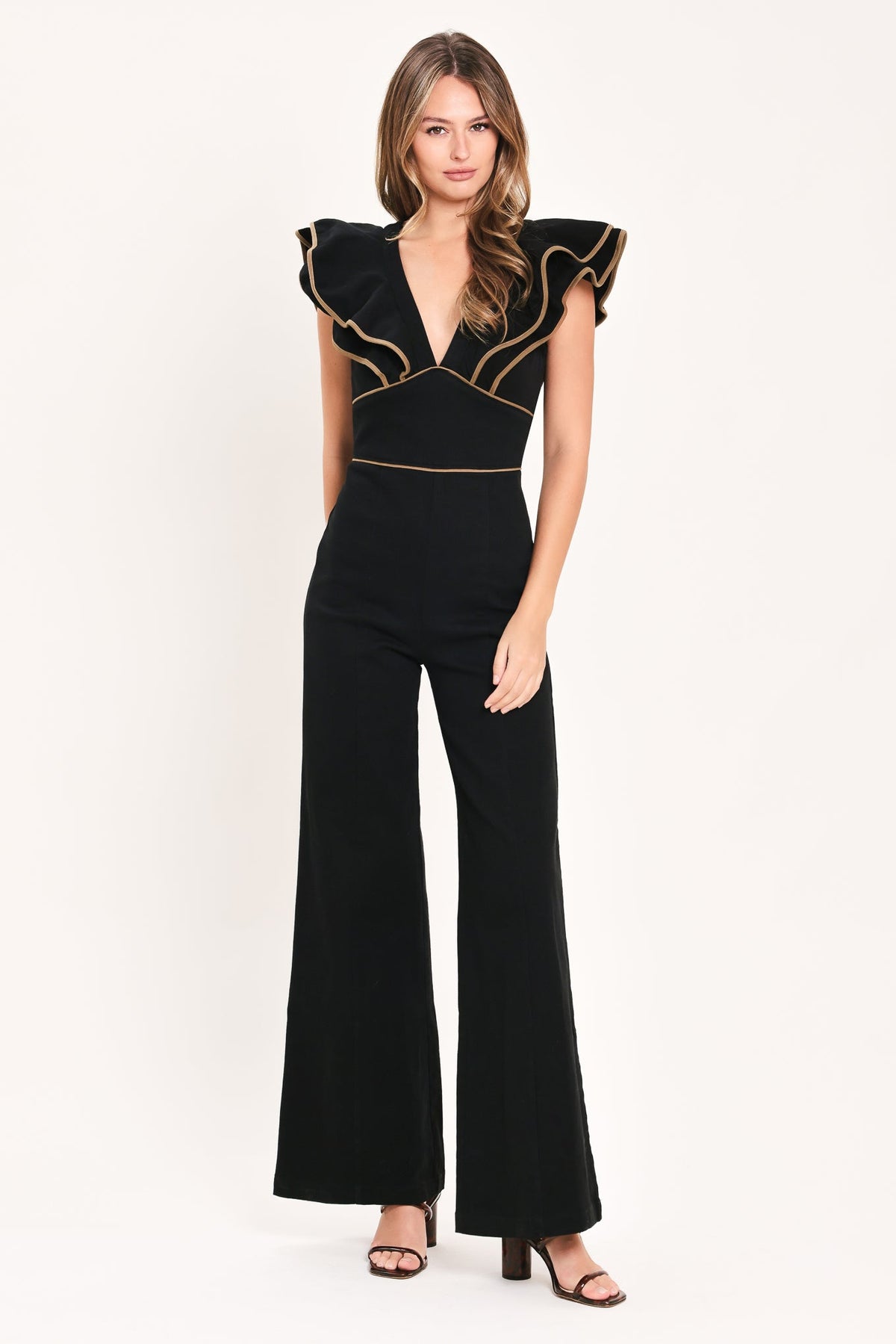 A woman modeling a black jumpsuit with dramatic ruffled cap sleeves, a deep V-neckline, and gold contrast piping details, set against a plain white studio background.