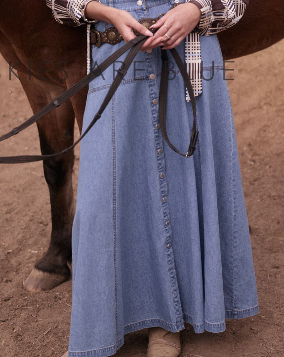 A person wearing a long light blue denim skirt with a button-front center and a dark belt, standing outdoors beside a horse on dusty ground. The person is holding the horse reins, and part of a patterned top is visible.
