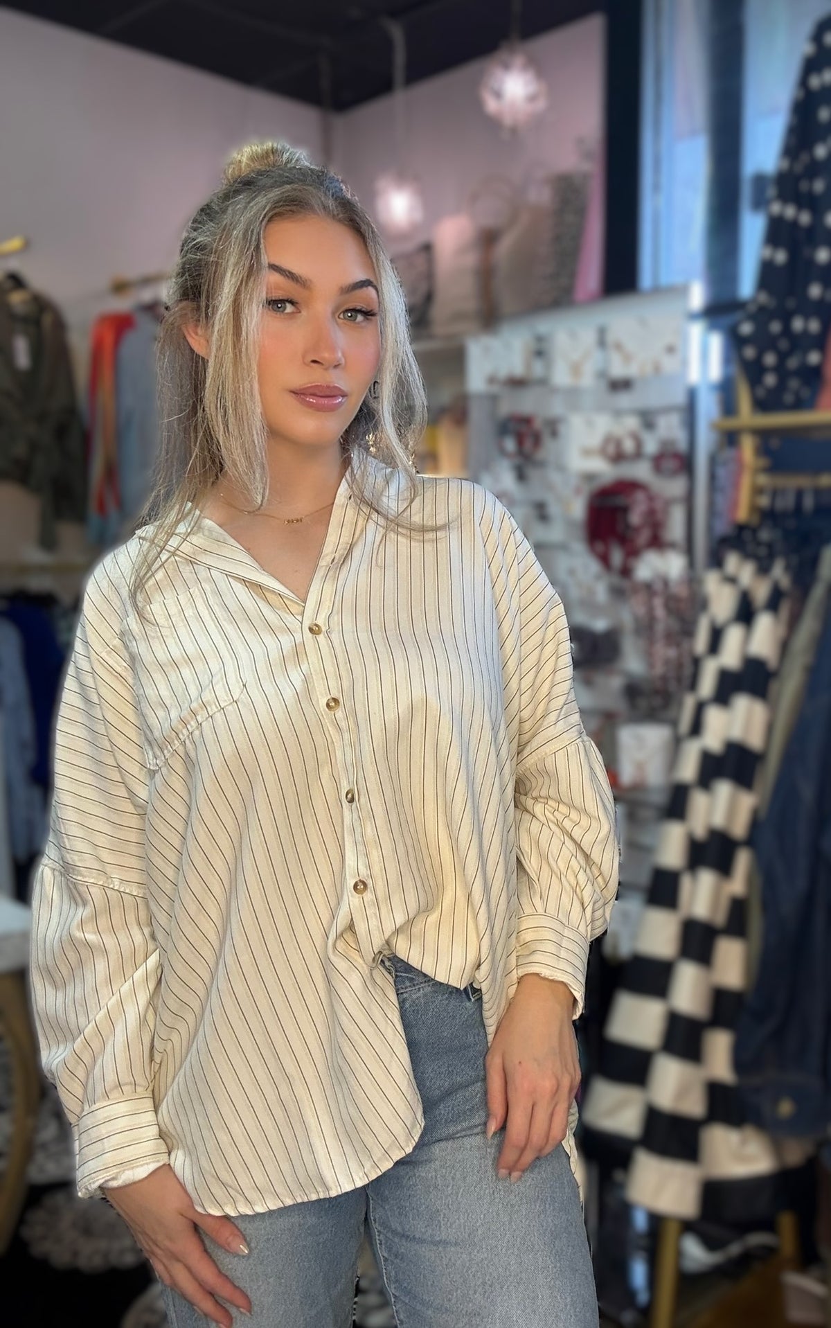 A woman with blonde hair in a bun wearing a cream and brown pinstriped button-down shirt and light-wash jeans, standing inside a clothing boutique.
