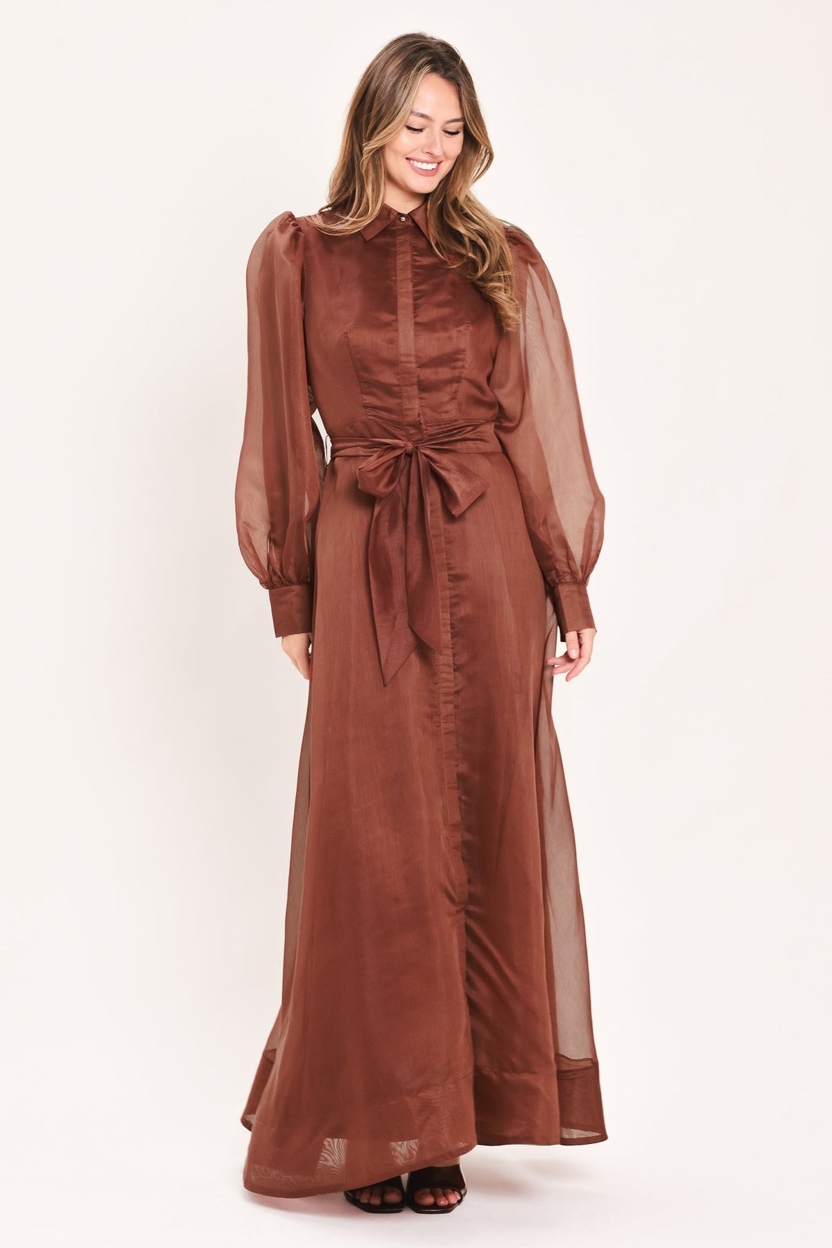 A woman modeling a rust-brown maxi dress with sheer balloon sleeves, a high ruffled collar, and a matching fabric tie belt at the waist, photographed against a plain light background.