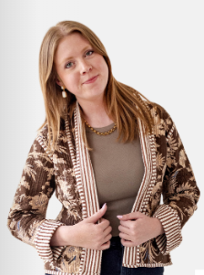 A woman with blonde hair wearing a brown and beige patterned cardigan over a taupe top, posing against a light gray background.