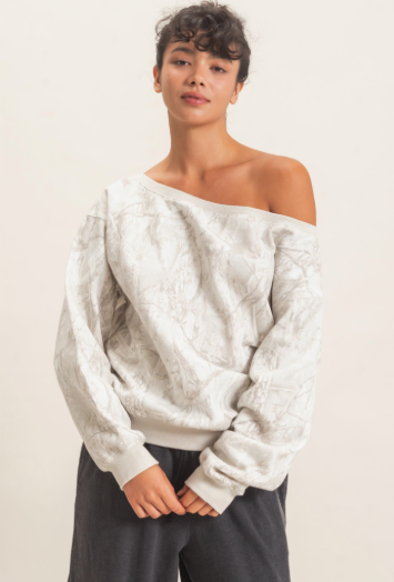 A woman modeling an off-the-shoulder knit sweater with a textured, tonal abstract pattern in off-white and light gray, paired with dark gray sweatpants against a plain light background.