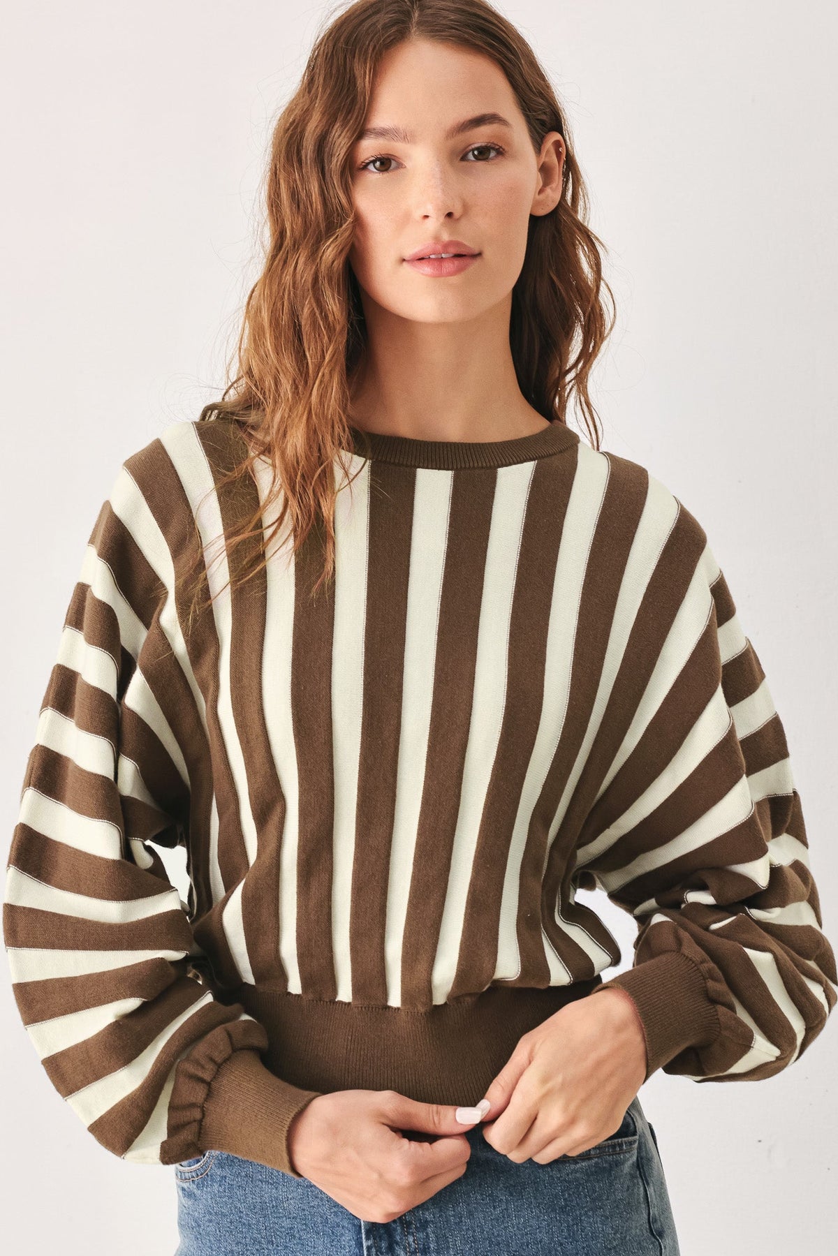 A woman wearing a brown and cream vertical striped long-sleeve knit top with a crew neck and elasticized hem, paired with blue jeans, against a light gray studio background.