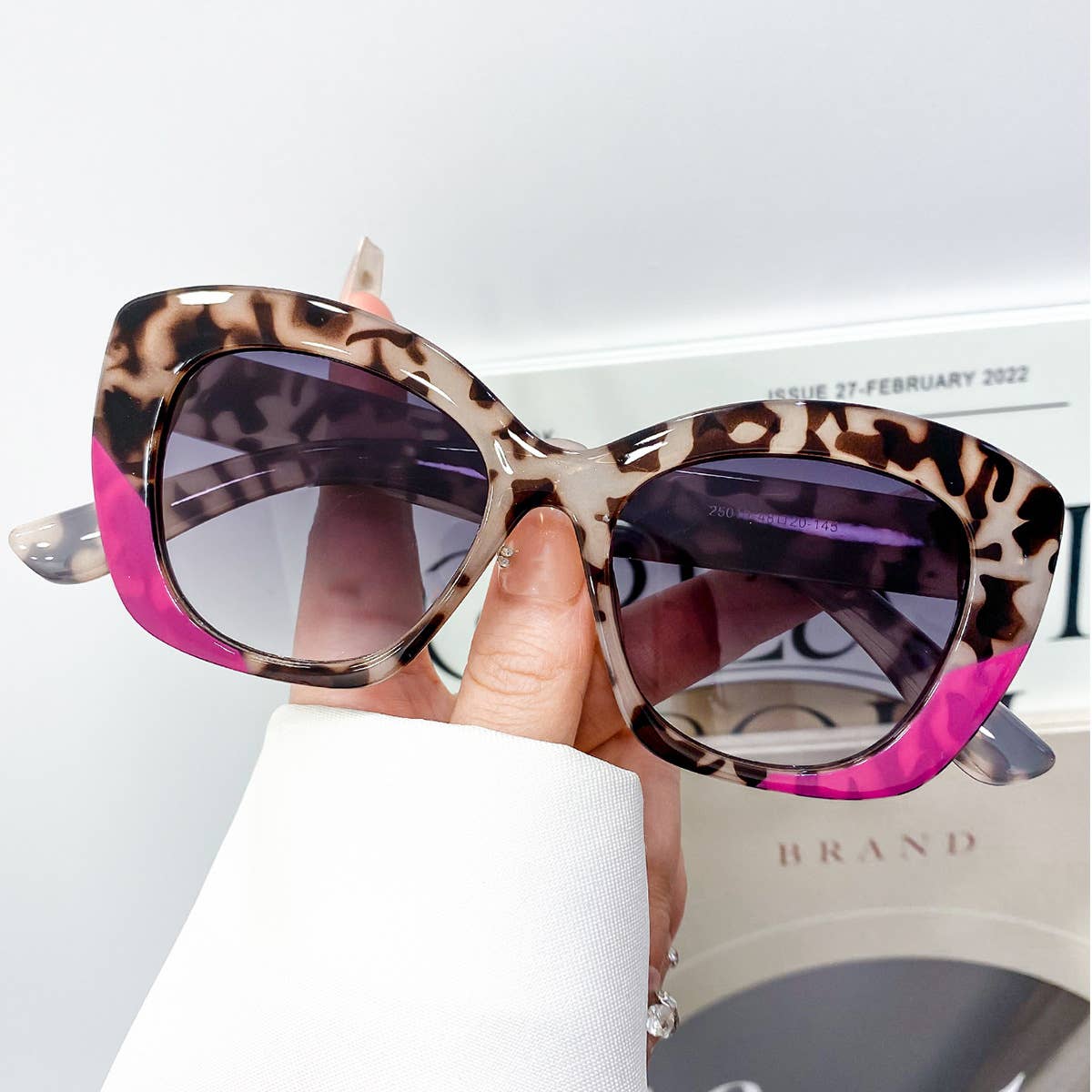Sunglasses with leopard print and pink frame held by a hand, with a magazine in the background.