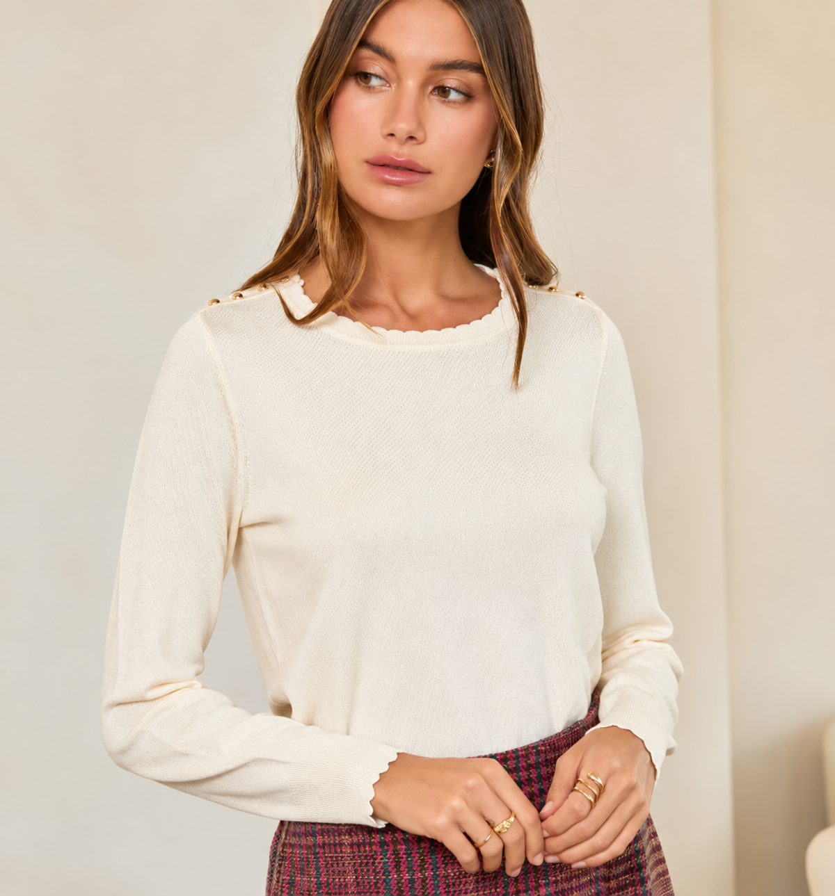A woman wearing a cream ribbed knit long-sleeve top with scalloped edges and small gold button details at the shoulders, paired with a red and blue plaid skirt.