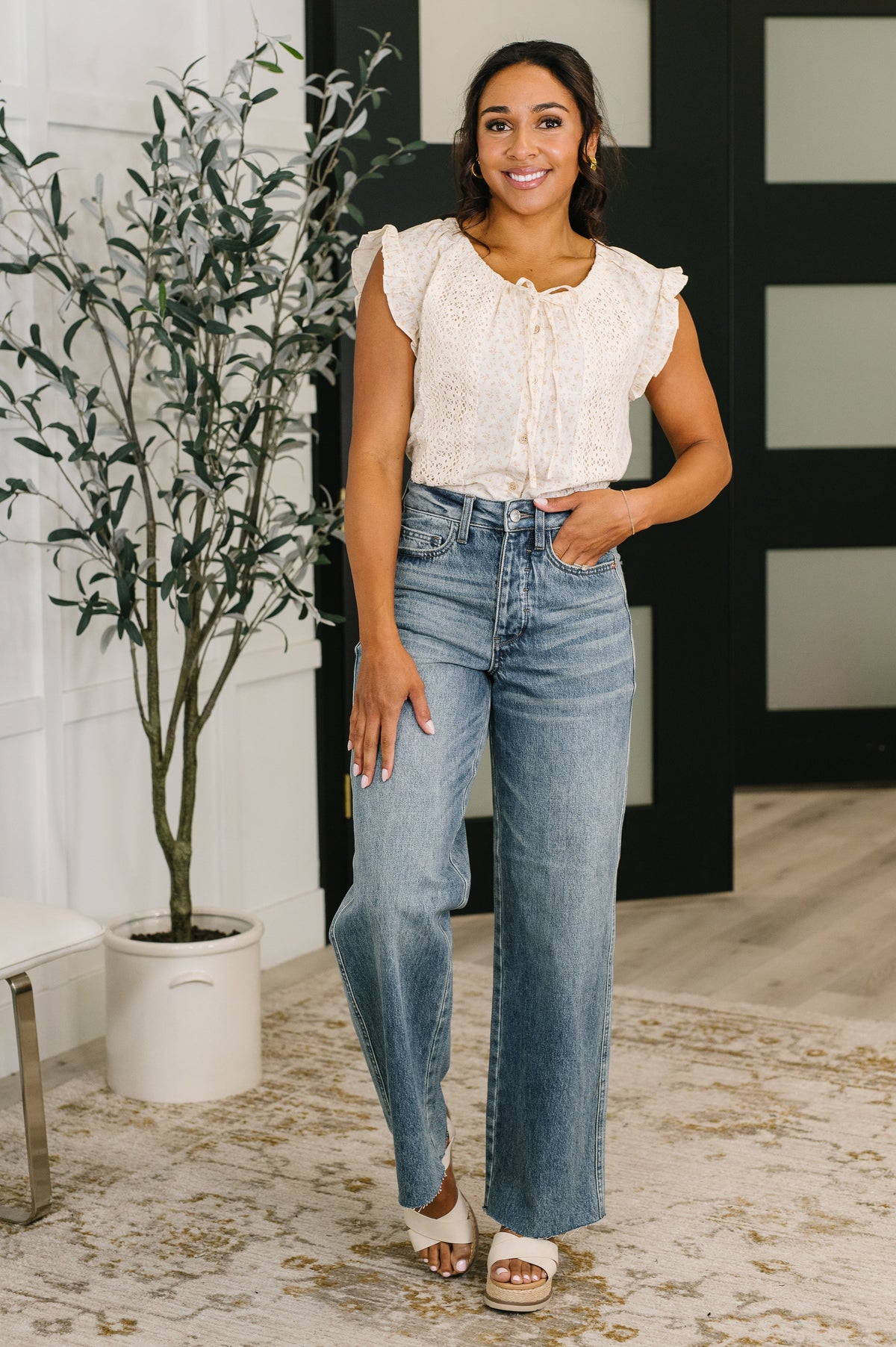 Woman wearing a white blouse and blue jeans standing indoors with a plant and decorative items in the background.