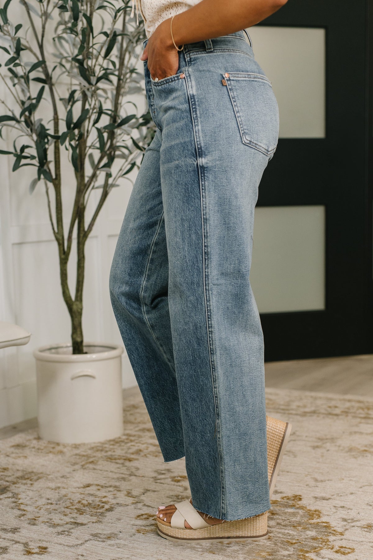 Person wearing light blue jeans standing indoors with a plant and wall decor in the background