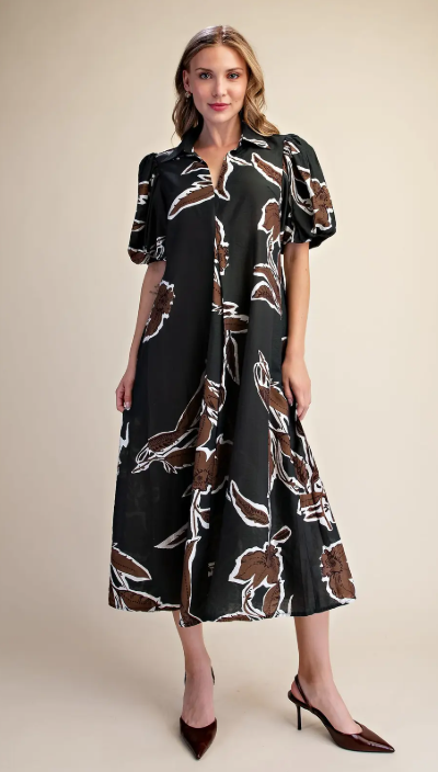 A woman modeling a black midi dress with a brown and white floral pattern, featuring short puff sleeves and a V-neckline, paired with dark brown pointed-toe heels against a solid beige studio background.