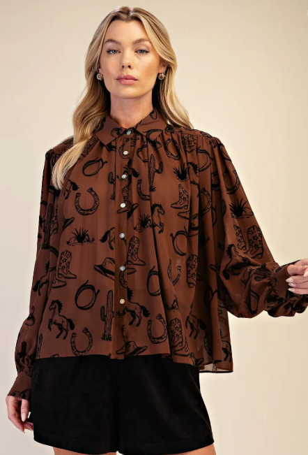 A woman modeling a brown long-sleeve button-up blouse with a black western-themed print featuring boots, horseshoes, cacti, and stars, paired with black shorts against a solid beige background.
