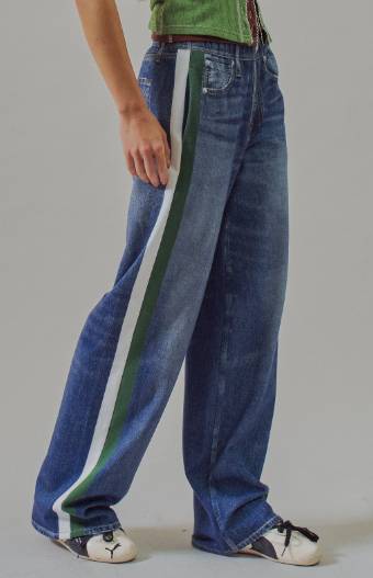 A person wearing wide-leg blue denim jeans with a bold green and white side stripe, paired with a green top and white sneakers, photographed against a plain light gray studio background.