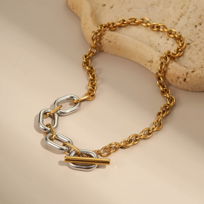 A gold and silver chain bracelet with a toggle clasp, styled on a neutral beige surface with a textured stone prop.
