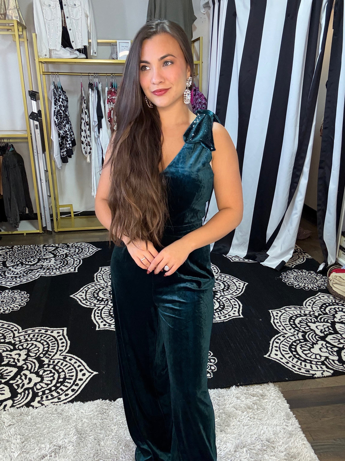 A woman standing in a retail setting wearing an emerald green, V-neck, wide-leg jumpsuit with sequin-embellished bows on the shoulders, paired with heels and statement earrings.