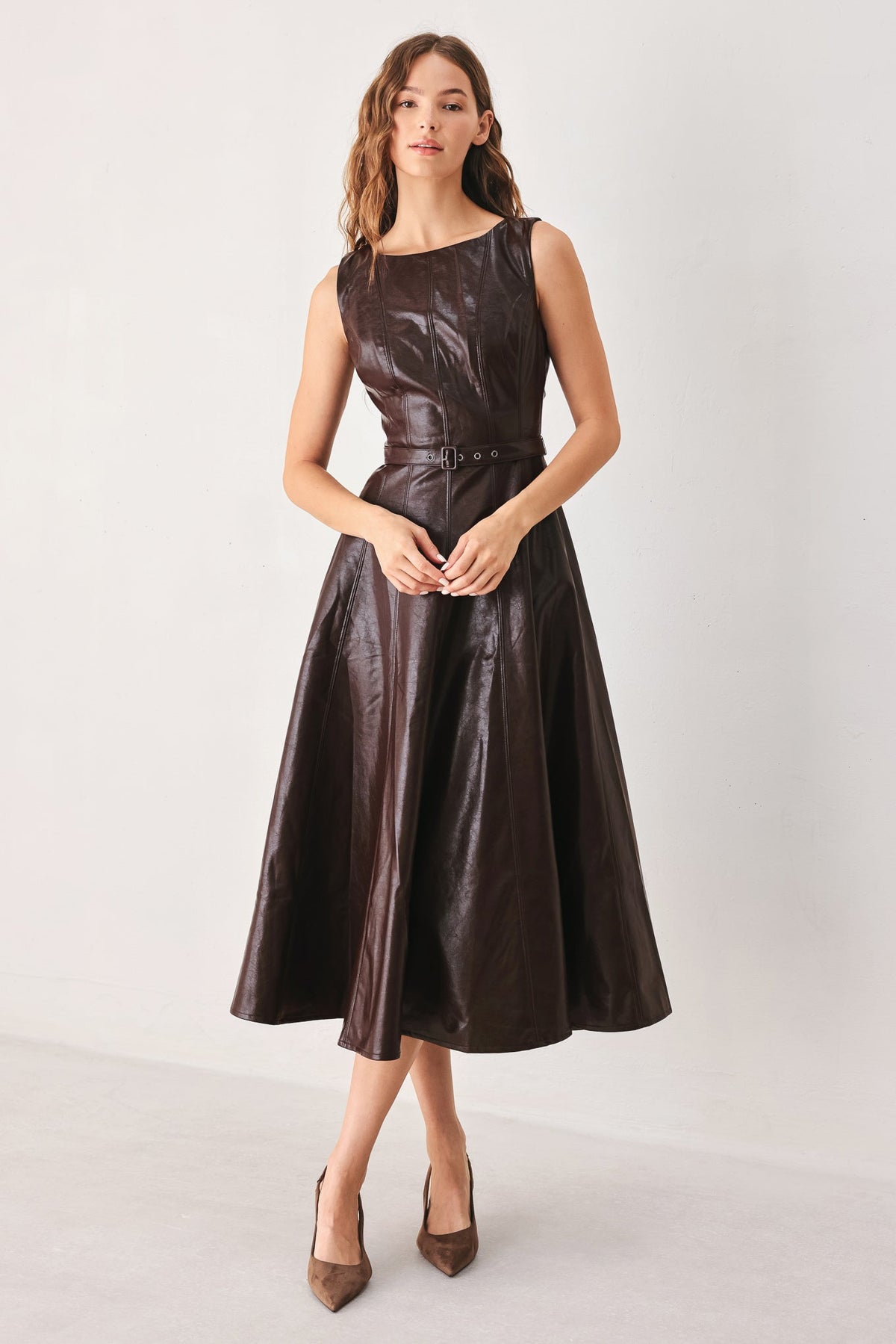 A woman modeling a sleeveless dark brown faux-leather midi dress with a cinched waist and flared skirt, paired with brown pointed-toe heels, against a plain light gray studio background.