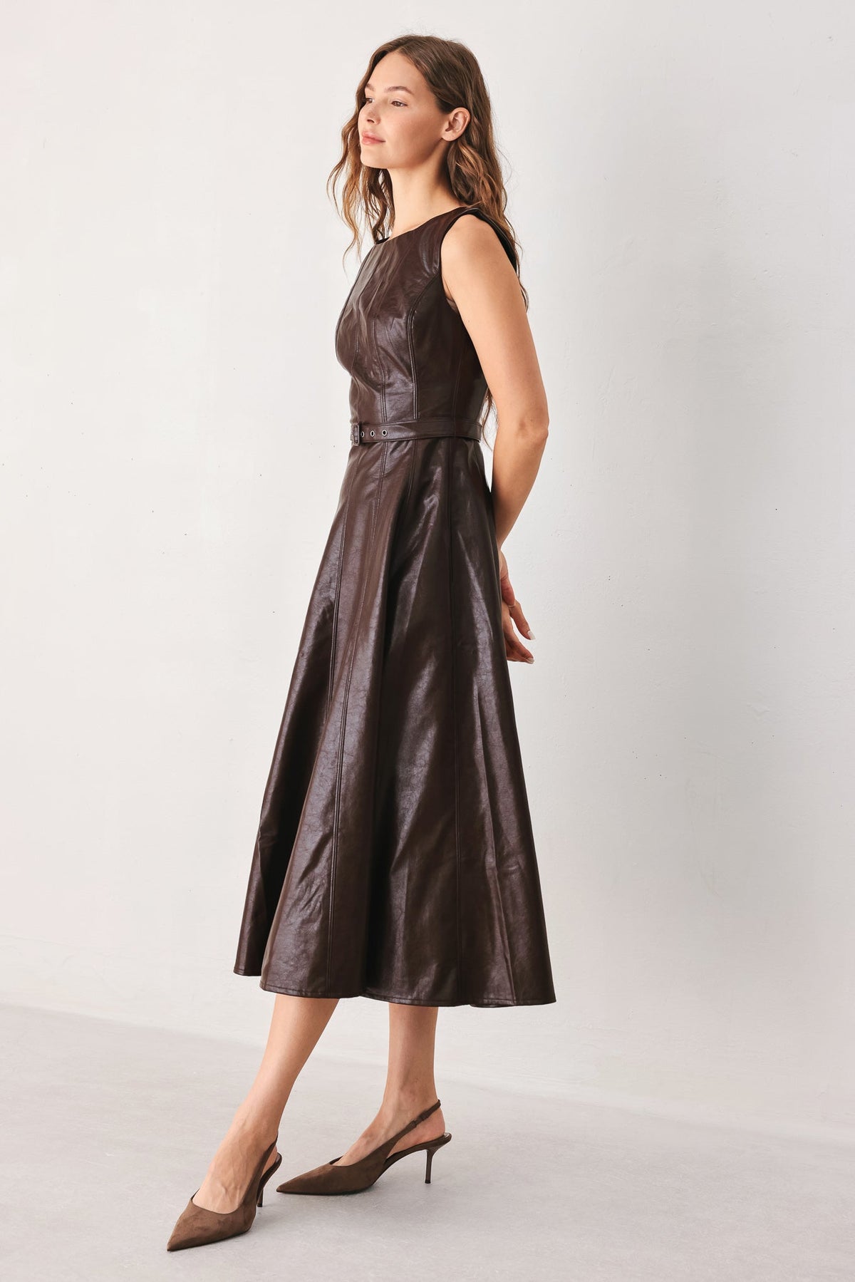 A woman modeling a sleeveless dark brown faux-leather midi dress with a structured bodice and flared skirt, paired with brown slingback heels, against a plain light gray studio background.