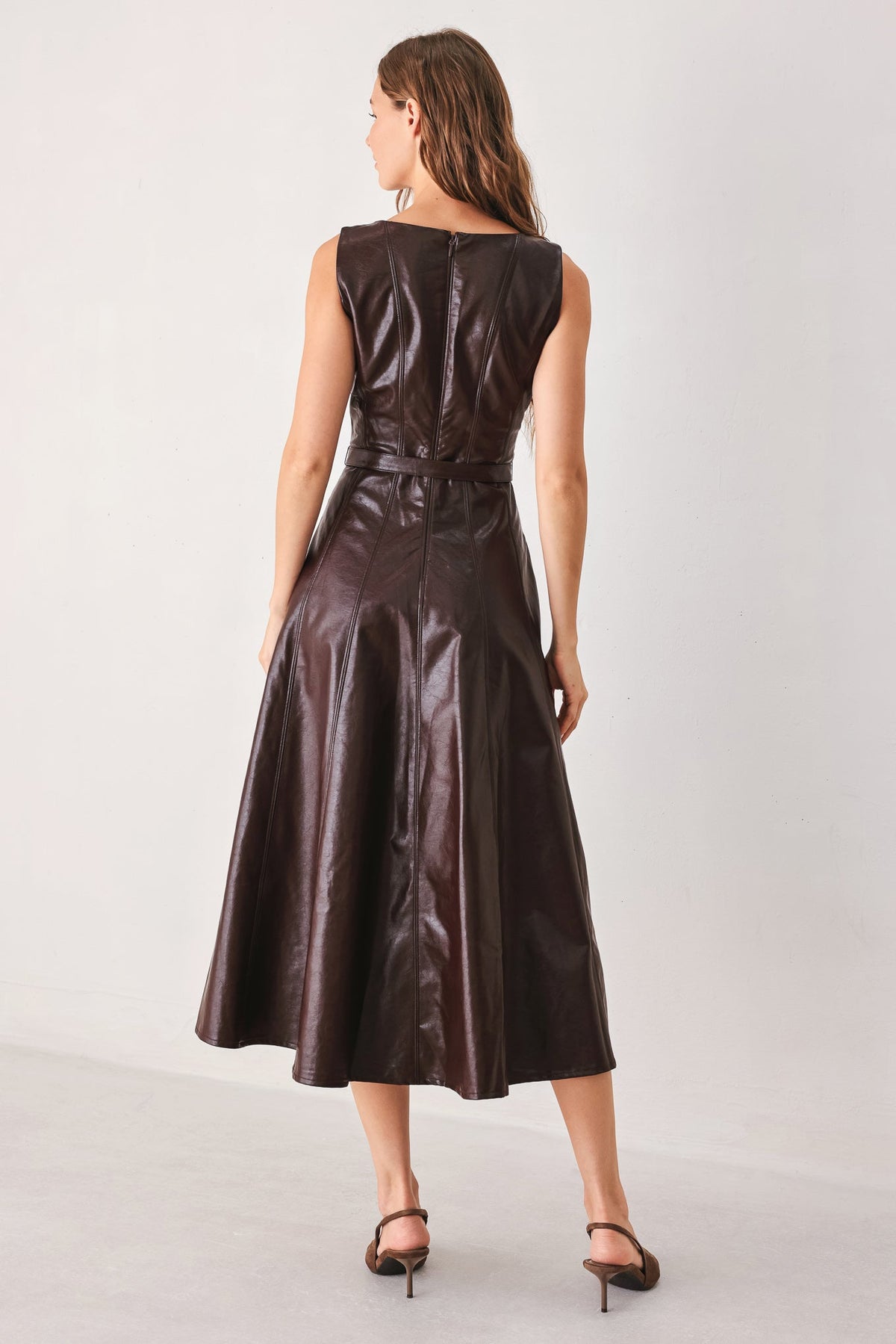 A woman viewed from the back wearing a sleeveless dark brown faux-leather midi dress with a fitted waist and flared skirt, paired with dark heeled sandals, against a plain light gray studio background.