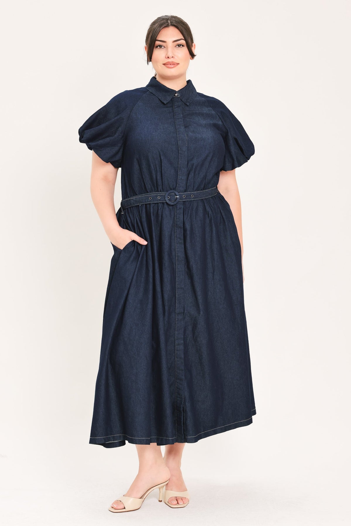 A woman modeling a dark navy blue denim midi dress with puff sleeves, a pointed collar, a matching fabric belt, and side pockets, set against a plain light background.