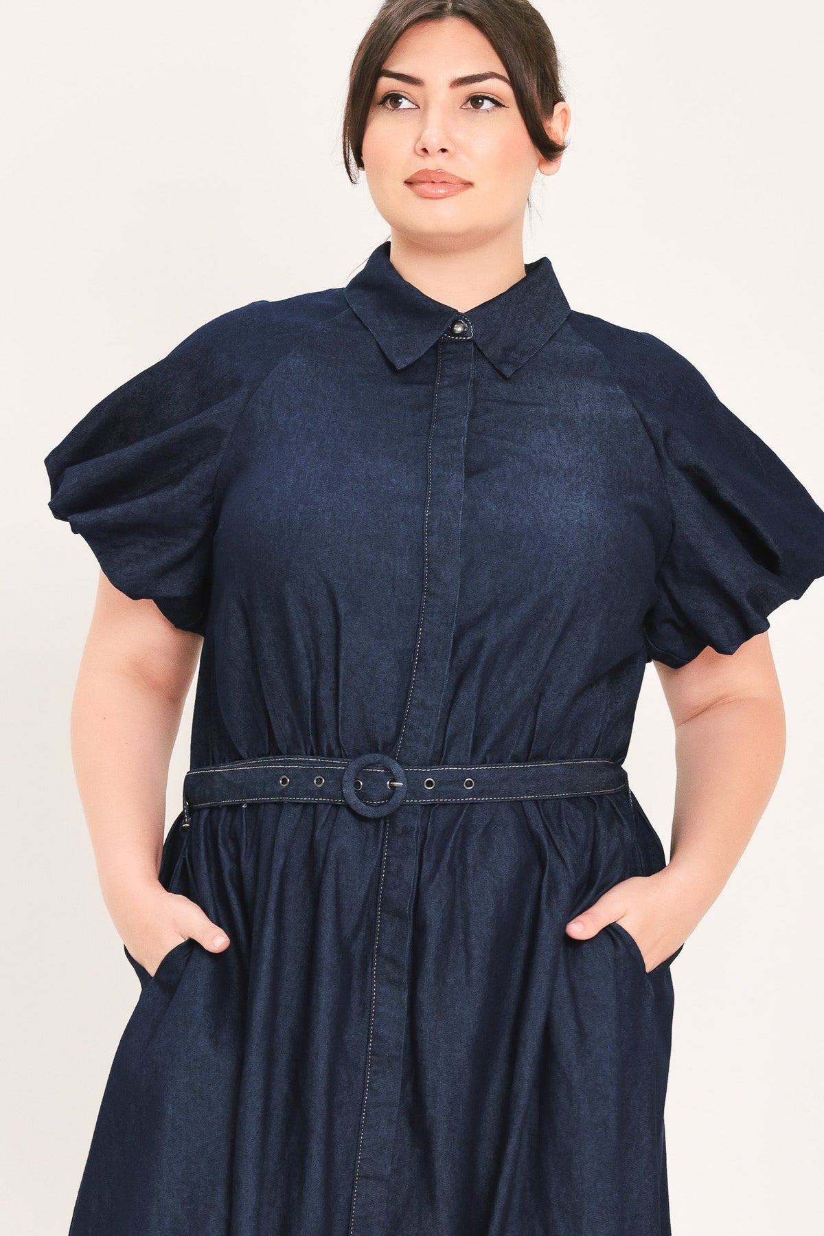 A woman wearing a dark navy blue chambray shirt dress with puff sleeves, a pointed collar, and a matching fabric belt with a round buckle, set against a plain off-white background.